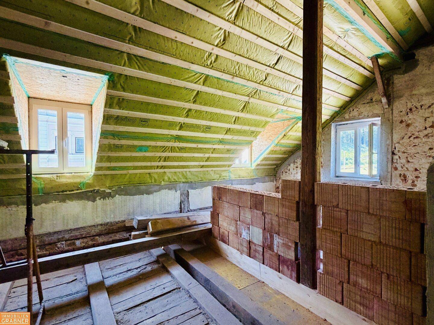 Dachgeschoss im Rohbau mit Isolierung, Holzbalken und neuen Fenstern, bereit für den Innenausbau.