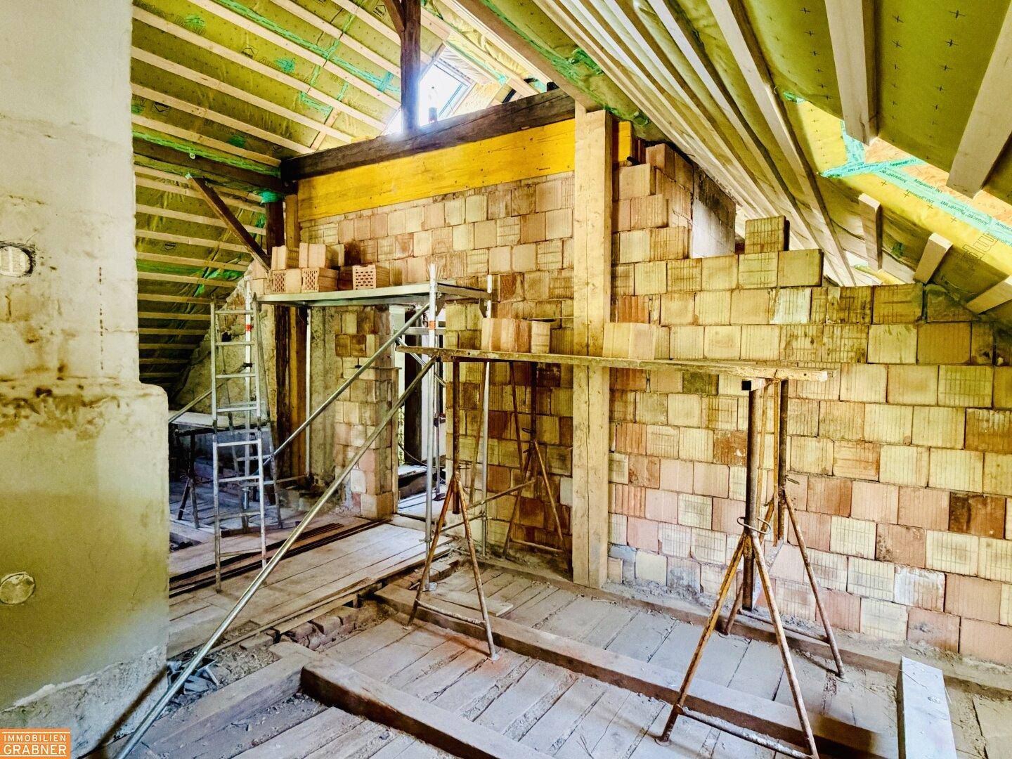 Dachgeschoss im Rohbau mit sichtbaren Ziegelwänden, Holzbalken und Isolierung, bereit für den Ausbau.