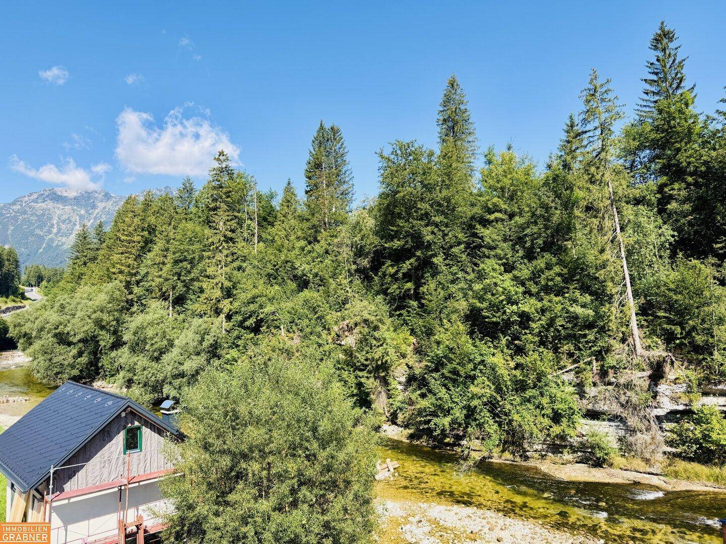 Malerischer Blick auf einen Fluss und dichte Wälder unter blauem Himmel, umgeben von Bergen.