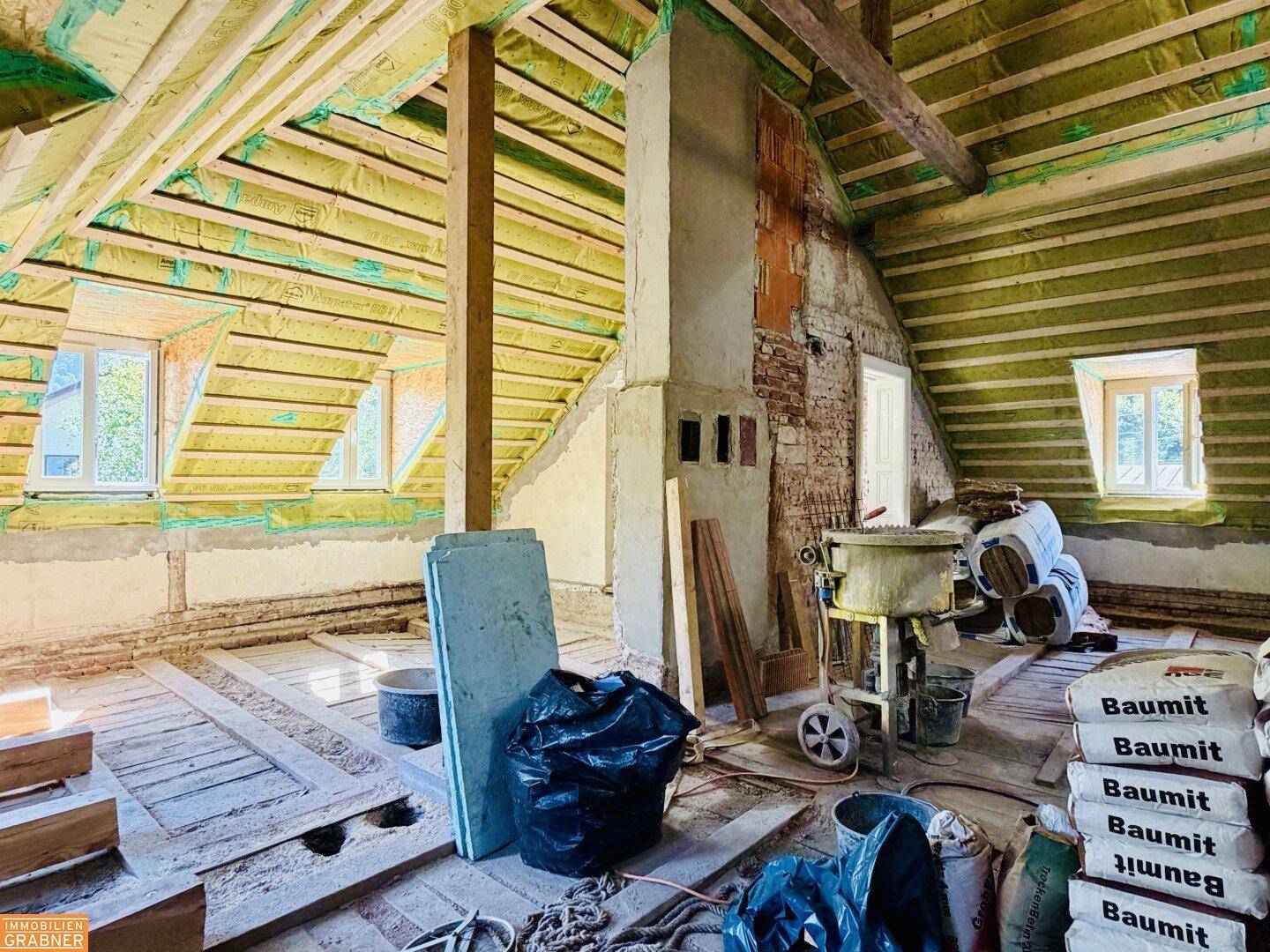 Dachgeschoss im Umbau mit Dämmung, Holzbalken, Kamin und Baumaterialien auf dem Boden.