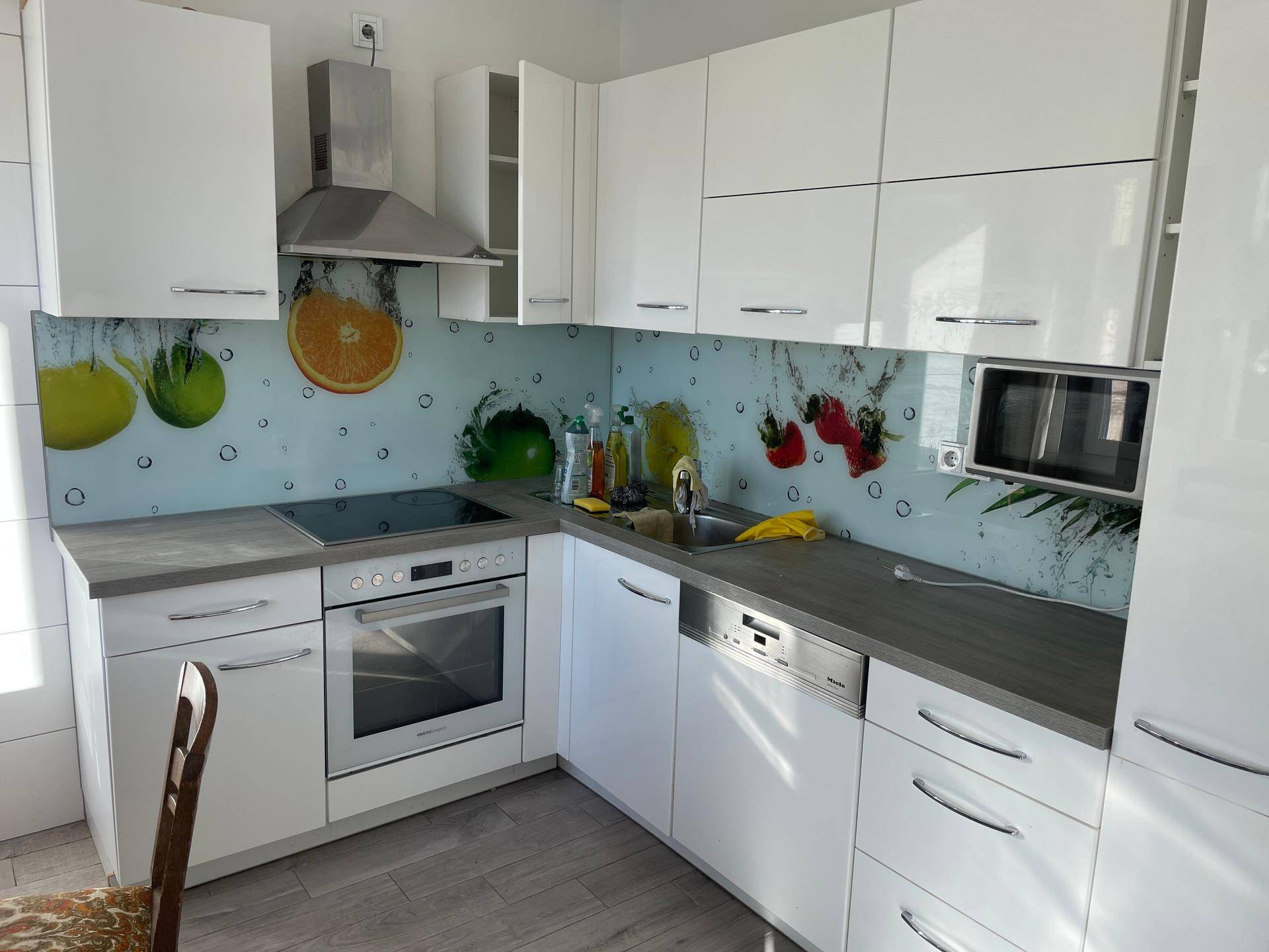 Moderne Einbauküche mit weißen Hochglanzschränken, grauen Arbeitsplatten und farbenfroher Rückwand.