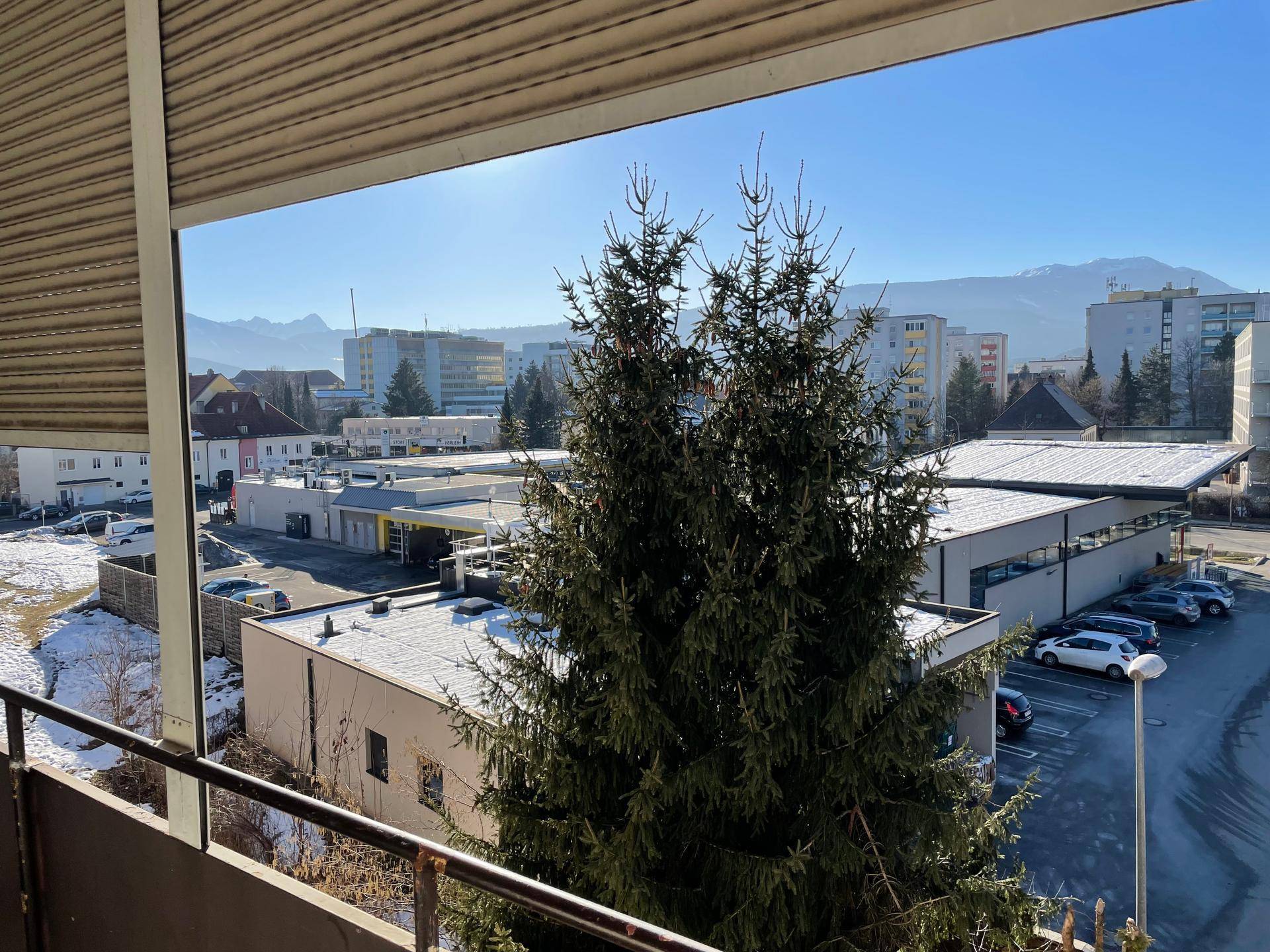 Sonniger Balkon mit Blick auf Bäume, städtische Bebauung und die beeindruckende Bergkulisse.