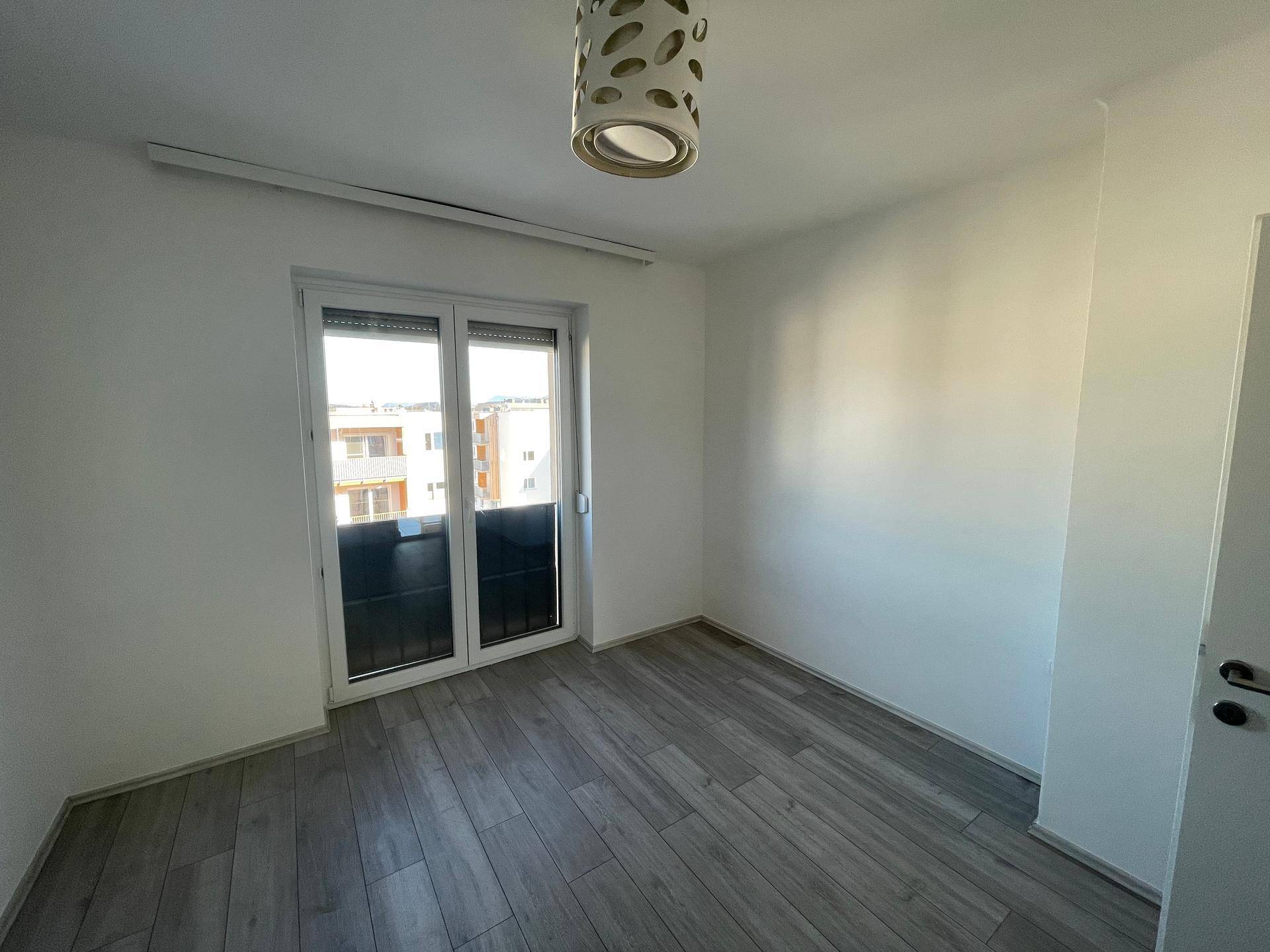 Schlafzimmer mit hellem Laminatboden, weißen Wänden und Zugang zu einem kleinen Balkon.
