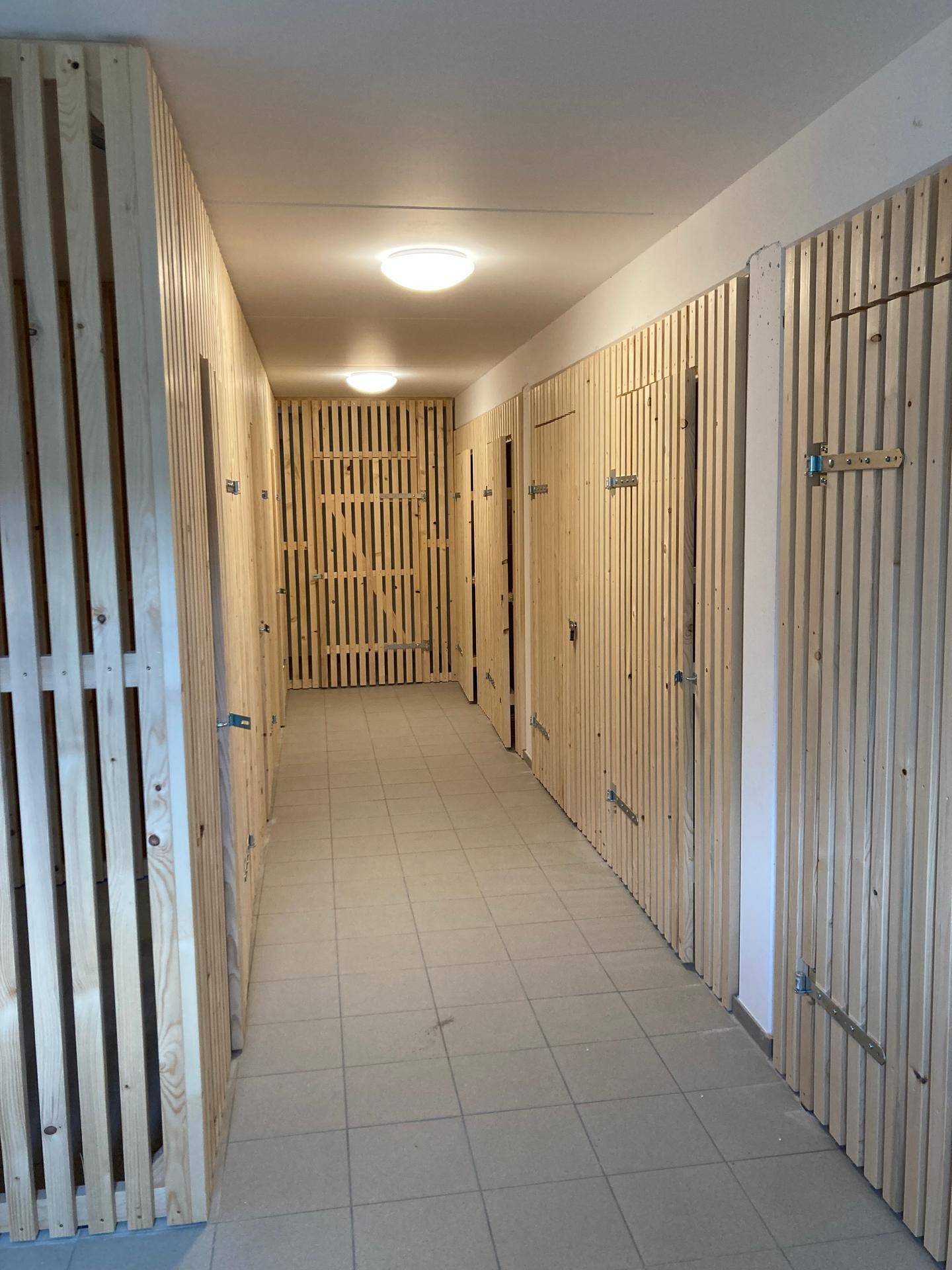Langer Flur im Keller mit individuellen Abstellräumen, die durch Holzlattenkonstruktionen abgetrennt sind.