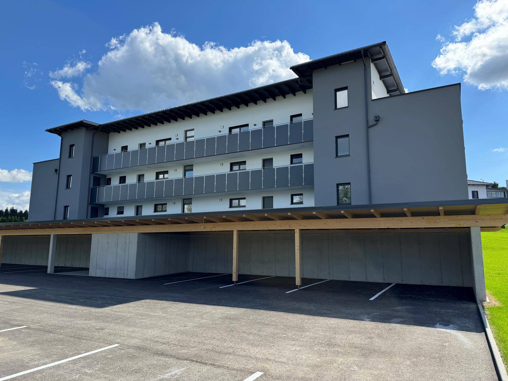 Ansicht des Neubaus mit Carports und gepflegter Grünfläche unter einem bewölkten Sommerhimmel.