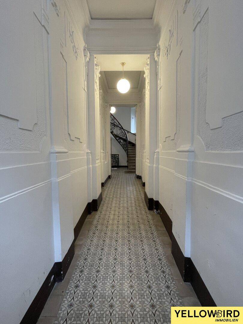 Langer, eleganter Flur mit dekorativen Wandpaneelen und einem gemusterten Fliesenboden, der zu einem Treppenhaus führt.