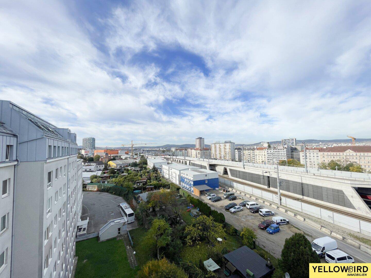 Weitläufiger Blick über die Stadt mit Gebäuden, Grünflächen und Bahngleisen unter bewölktem Himmel.