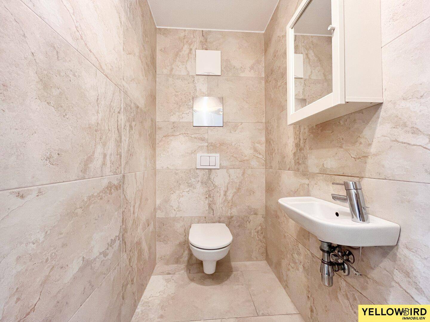 Separates WC mit beigefarbenen Fliesen, wandhängender Toilette und kleinem Handwaschbecken.