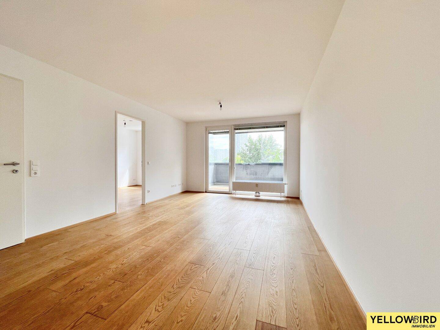 Helles, leeres Wohnzimmer mit Parkettboden und Zugang zu einem Balkon oder einer Loggia.