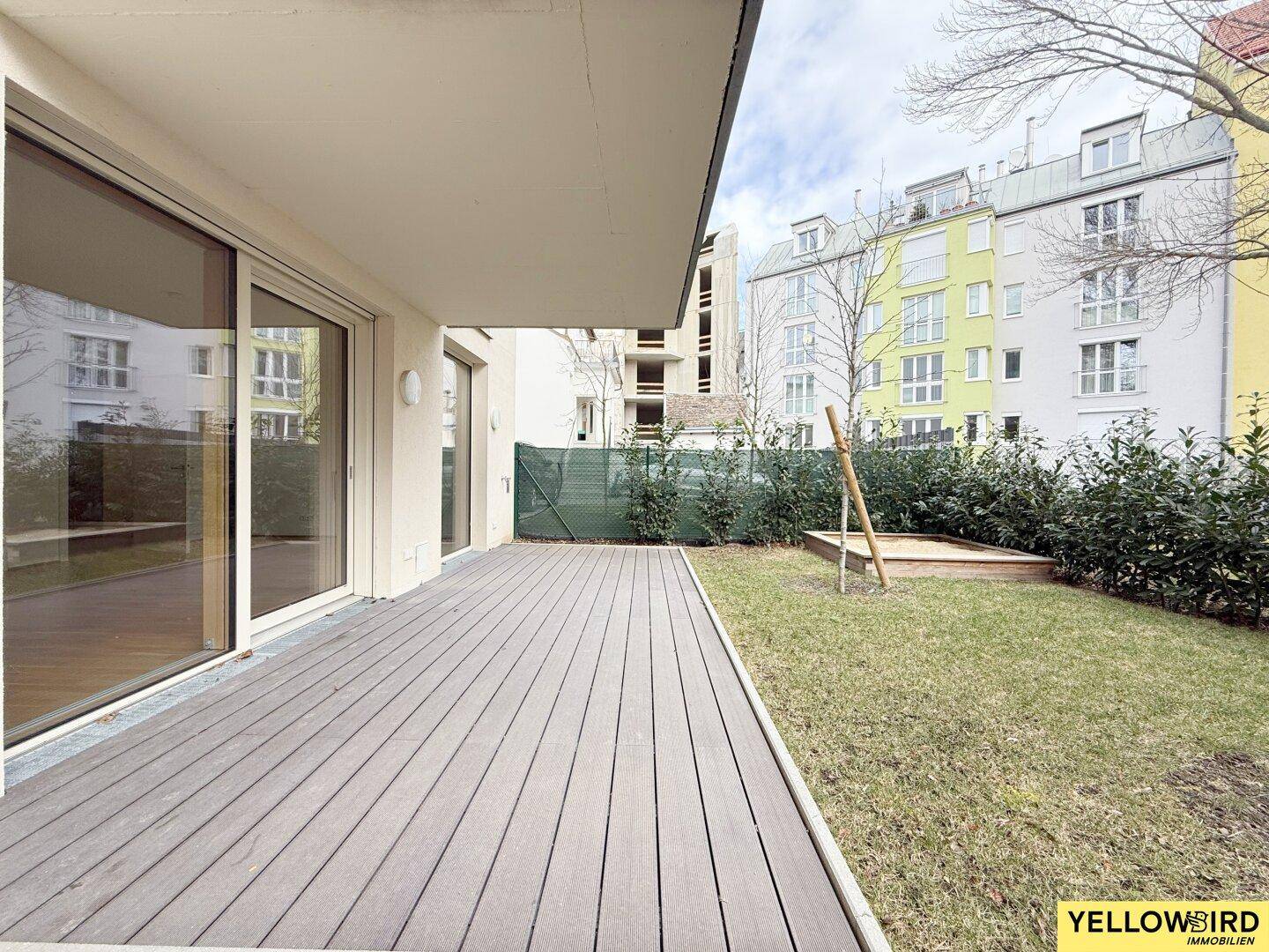 Geräumige Terrasse mit Holzbelag, kleiner Rasenfläche und Sandkasten, umgeben von Wohngebäuden.