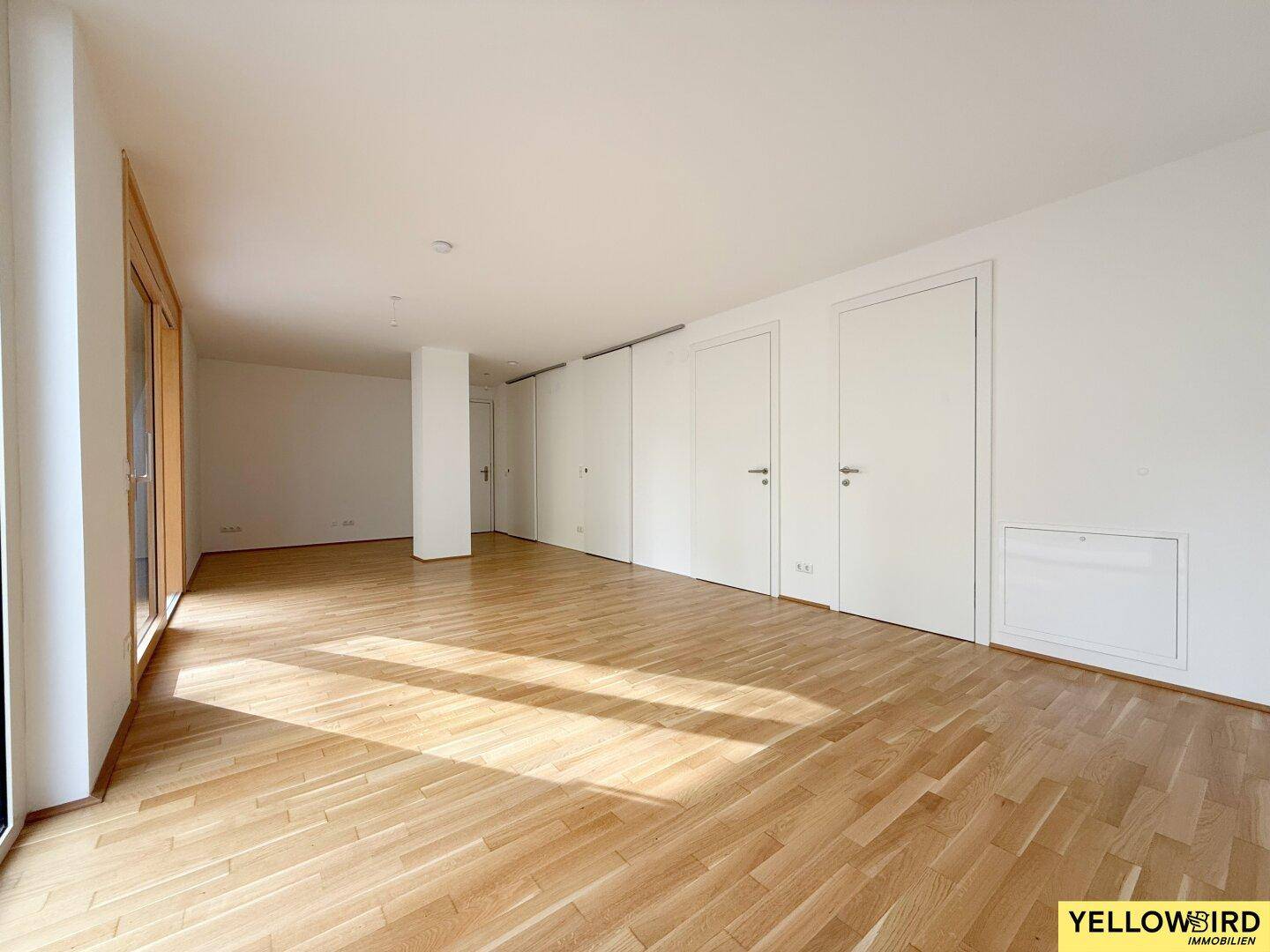 Geräumiger, leerer Wohnbereich mit hellem Holzboden und viel natürlichem Lichteinfall durch die Fenster.