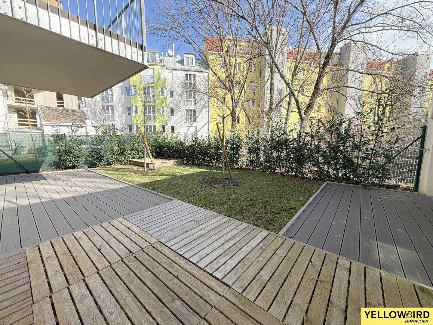 Außenbereich mit Holzterrasse und Rasenfläche, umgeben von Bäumen und anderen Gebäuden.