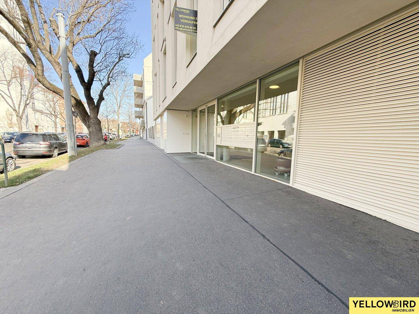 Moderne Gebäudefassade mit großen Fenstern und einem Gehweg, der an der Straße entlangführt.