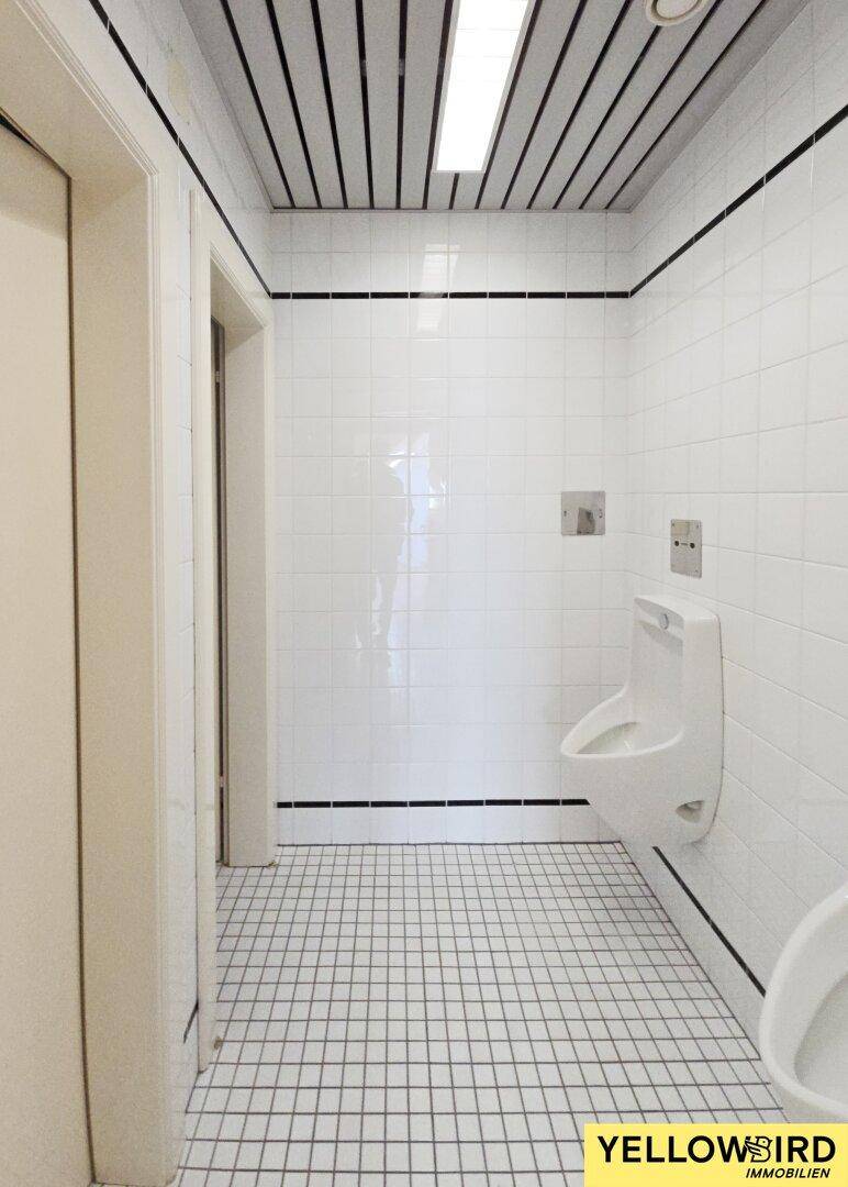 Gefliestes Gäste-WC mit Urinal, weißen Fliesen und schwarzen Fugen, sowie einer Deckenbeleuchtung.
