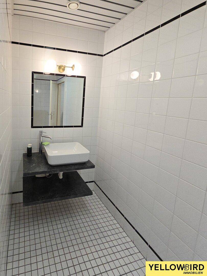 Modern gefliestes Gäste-WC mit weißem Waschbecken, schwarzem Akzent und einem Spiegel über dem Waschtisch.