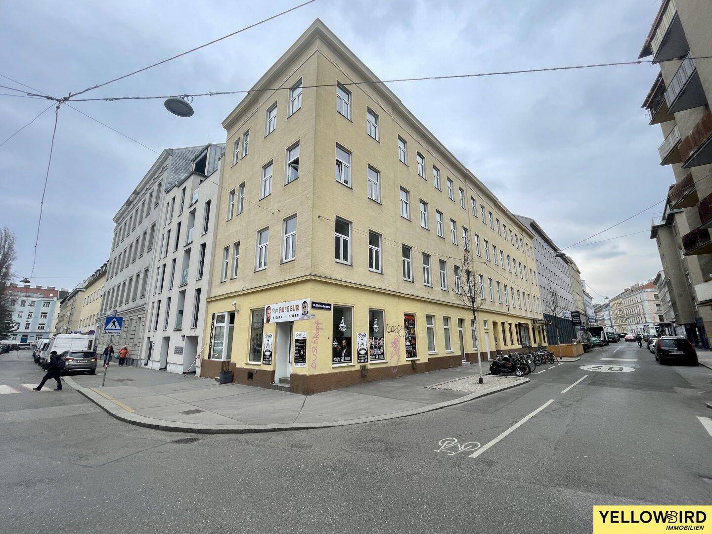 Gelbes Eckgebäude mit Geschäften im Erdgeschoss und einer belebten Straße davor.