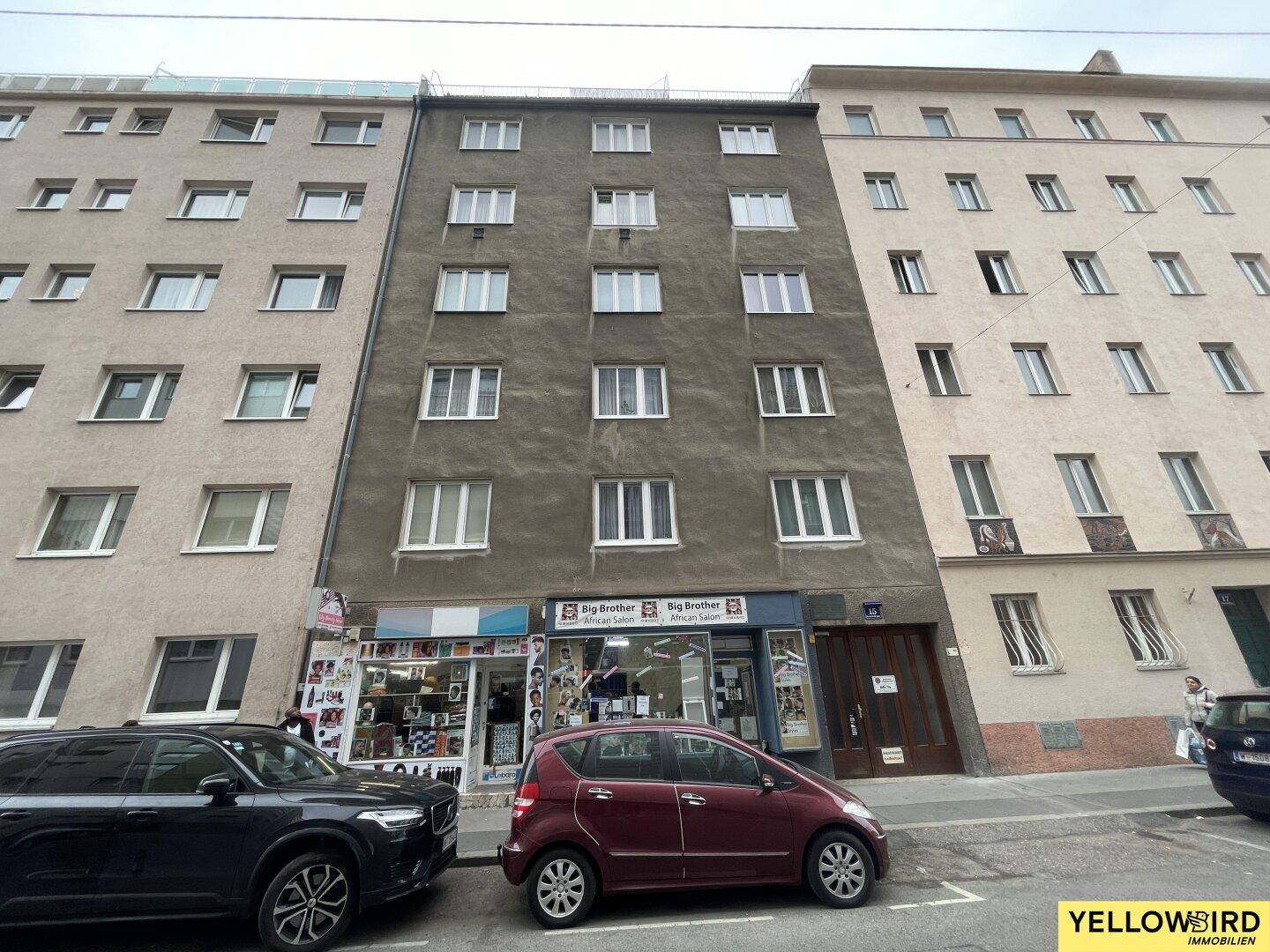 Graue Fassade eines Wohngebäudes mit Geschäften im Erdgeschoss und parkenden Autos davor.