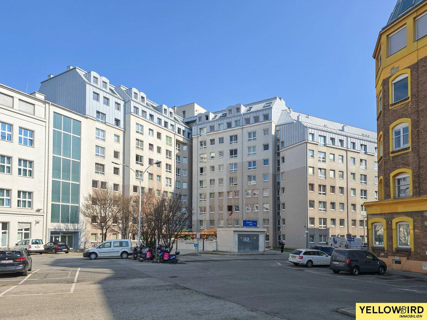 Großer Wohnkomplex mit mehreren Gebäuden, Parkplätzen und Mansarddächern unter blauem Himmel.