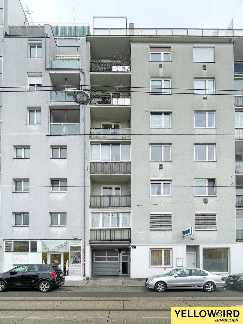 Ansicht der grauen Fassade eines Mehrfamilienhauses mit mehreren Balkonen und Geschäften im Erdgeschoss.