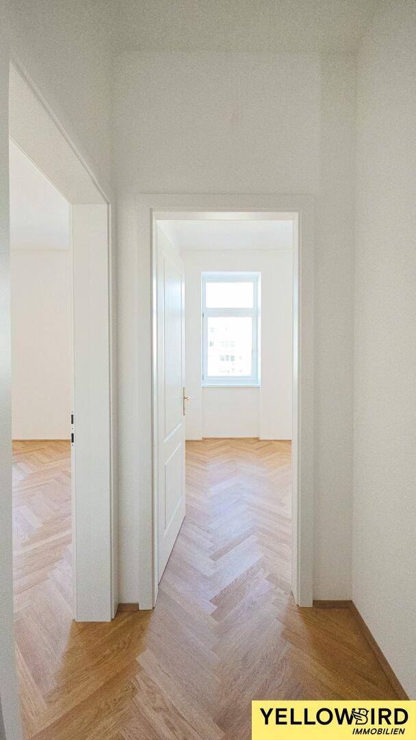 Blick vom Flur in einen hellen, leeren Raum mit Fischgrätparkett und einem Fenster.