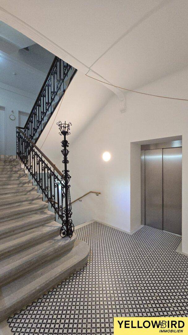 Heller Treppenaufgang mit schmiedeeisernem Geländer und modernem Aufzug im Gebäude.