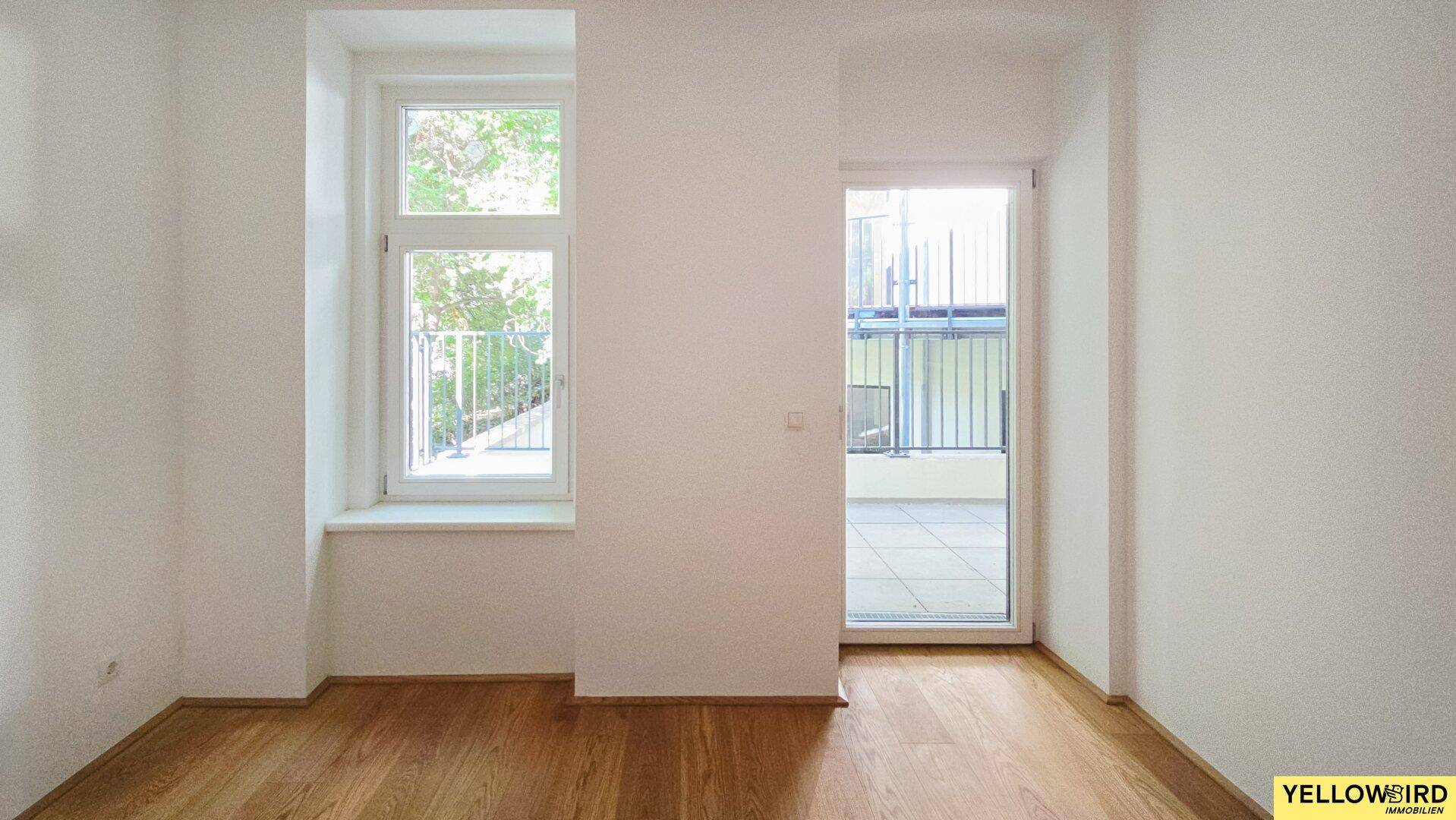 Leeres Zimmer mit Holzboden, großem Fenster und Zugang zu einem Balkon.