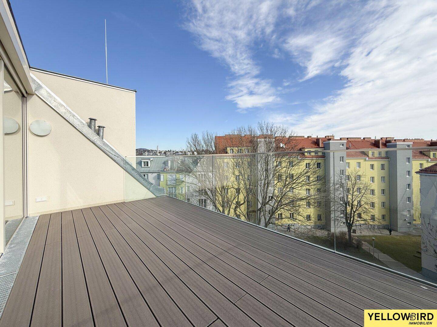Weitläufige Dachterrasse mit Holzboden und Glasgeländer, umgeben von städtischer Bebauung.