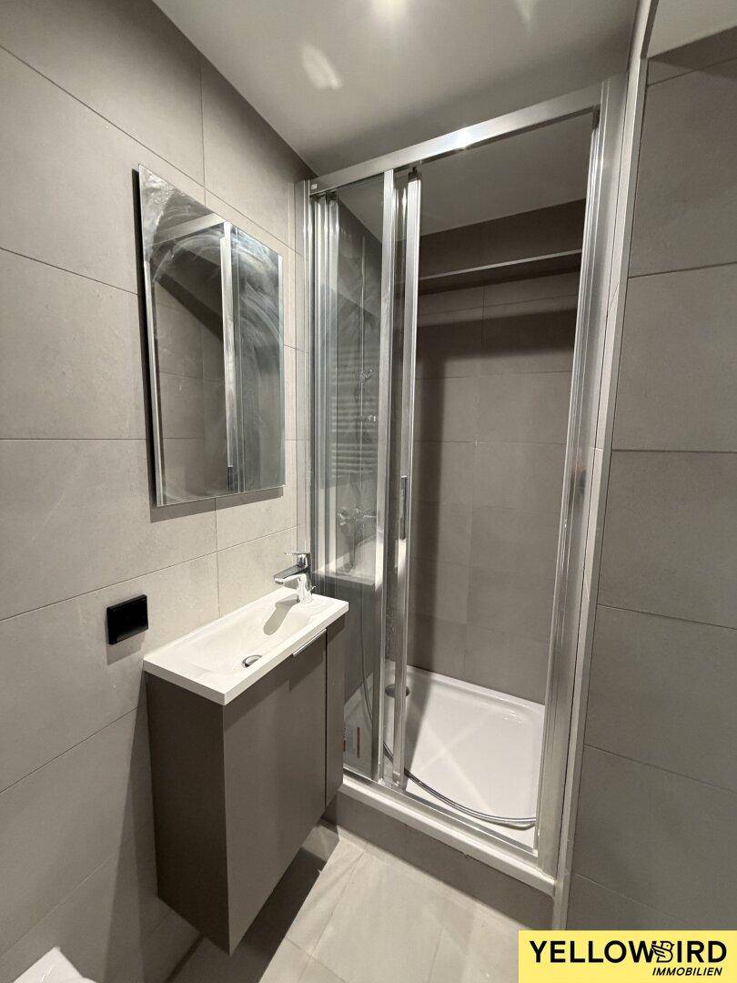Modernes Badezimmer mit grauen Fliesen, Duschkabine, Waschtisch und Spiegel, in gutem Zustand.