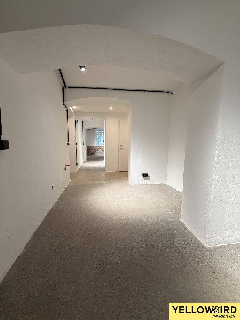 Weiterer Blick in den hellen Flur mit Betonboden und weißen Wänden, der in offene Räume mündet.