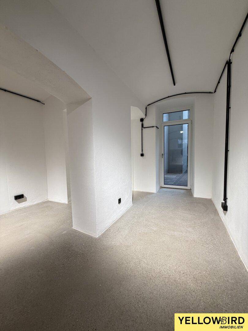 Heller Flur mit Betonboden, weißen Wänden und einer Glastür, die zu einem Außenbereich führt.