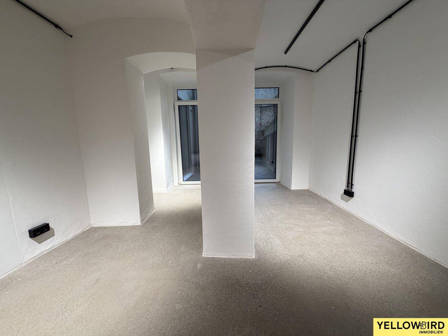 Heller Innenraum mit Betonboden, weißen Wänden, Säule und zwei Glastüren zum Außenbereich.