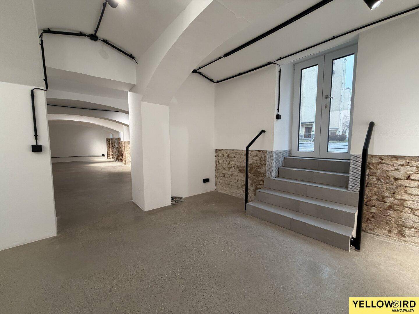 Heller Gewerberaum mit Gewölbedecken, Betonboden und Treppe, die zu einer Außentür führt.