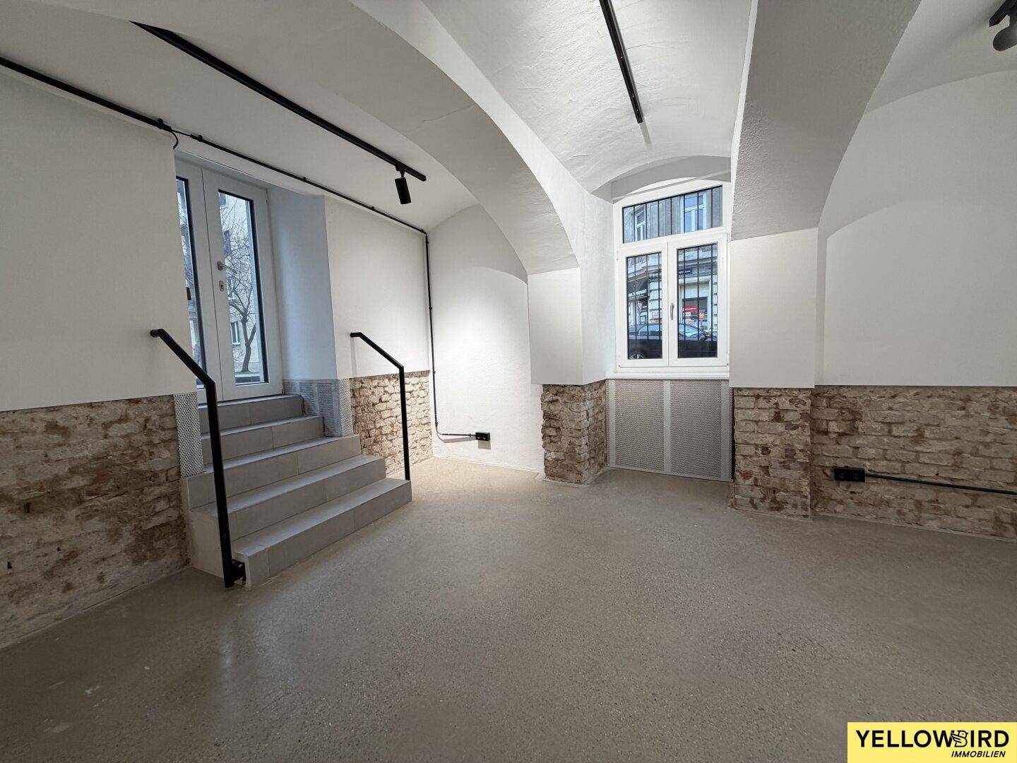 Großer offener Raum mit Gewölbedecken, Betonboden, freigelegten Ziegeln und Treppe zu einer Glastür.