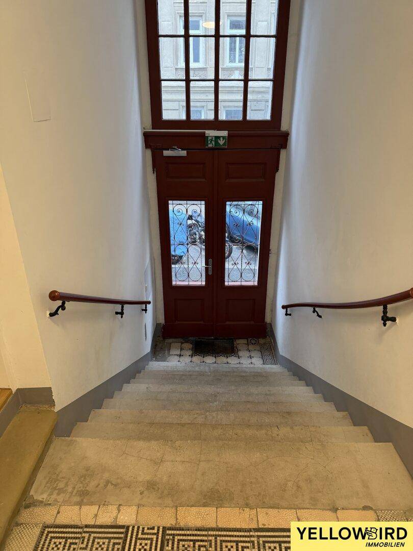 Historisches Treppenhaus mit Mosaikboden, Holzgeländer und einer großen Holztür mit Buntglasfenstern.