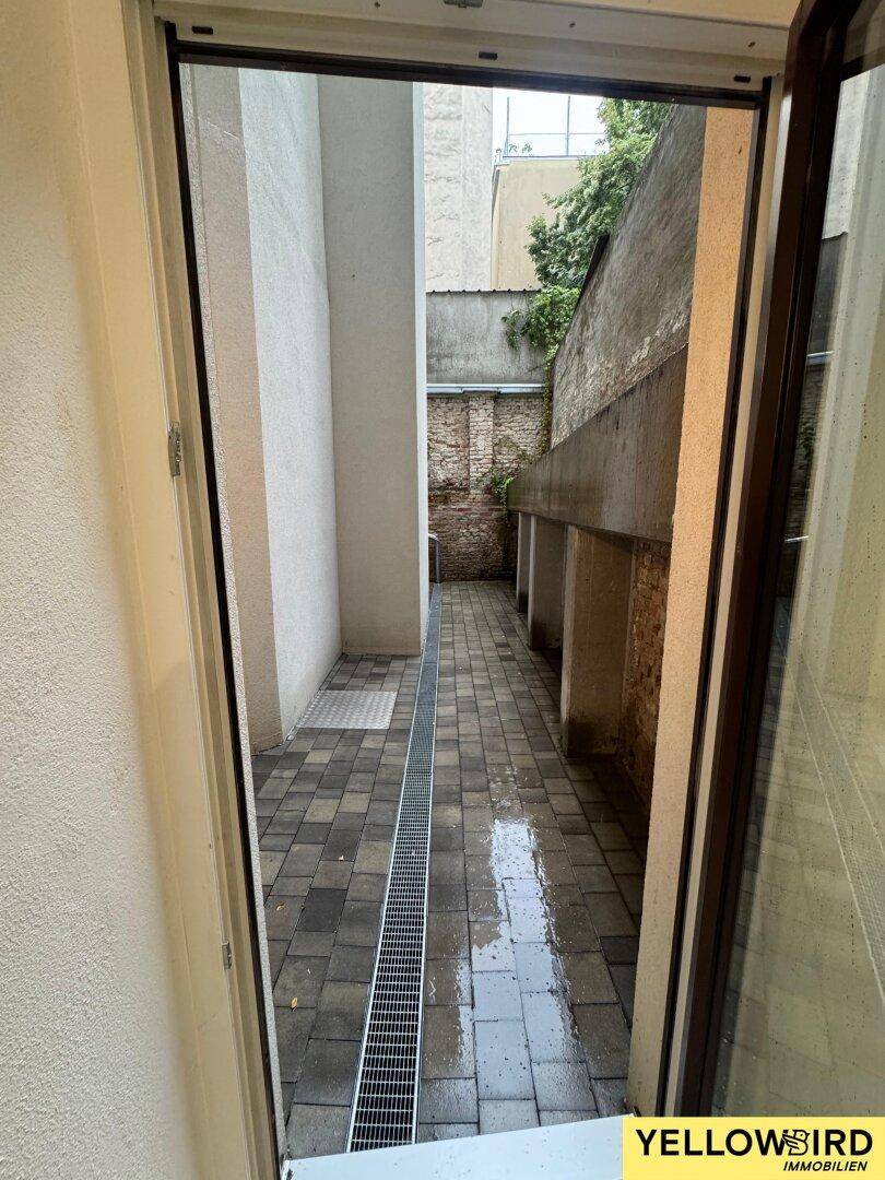 Blick aus einer Tür in einen schmalen, gepflasterten Innenhof mit Entwässerungsrinne.