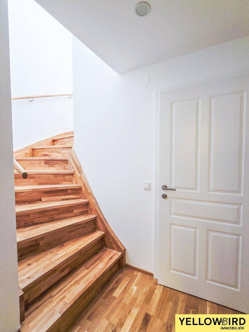 Heller Treppenaufgang mit Holzstufen und weiß gestrichenen Wänden.