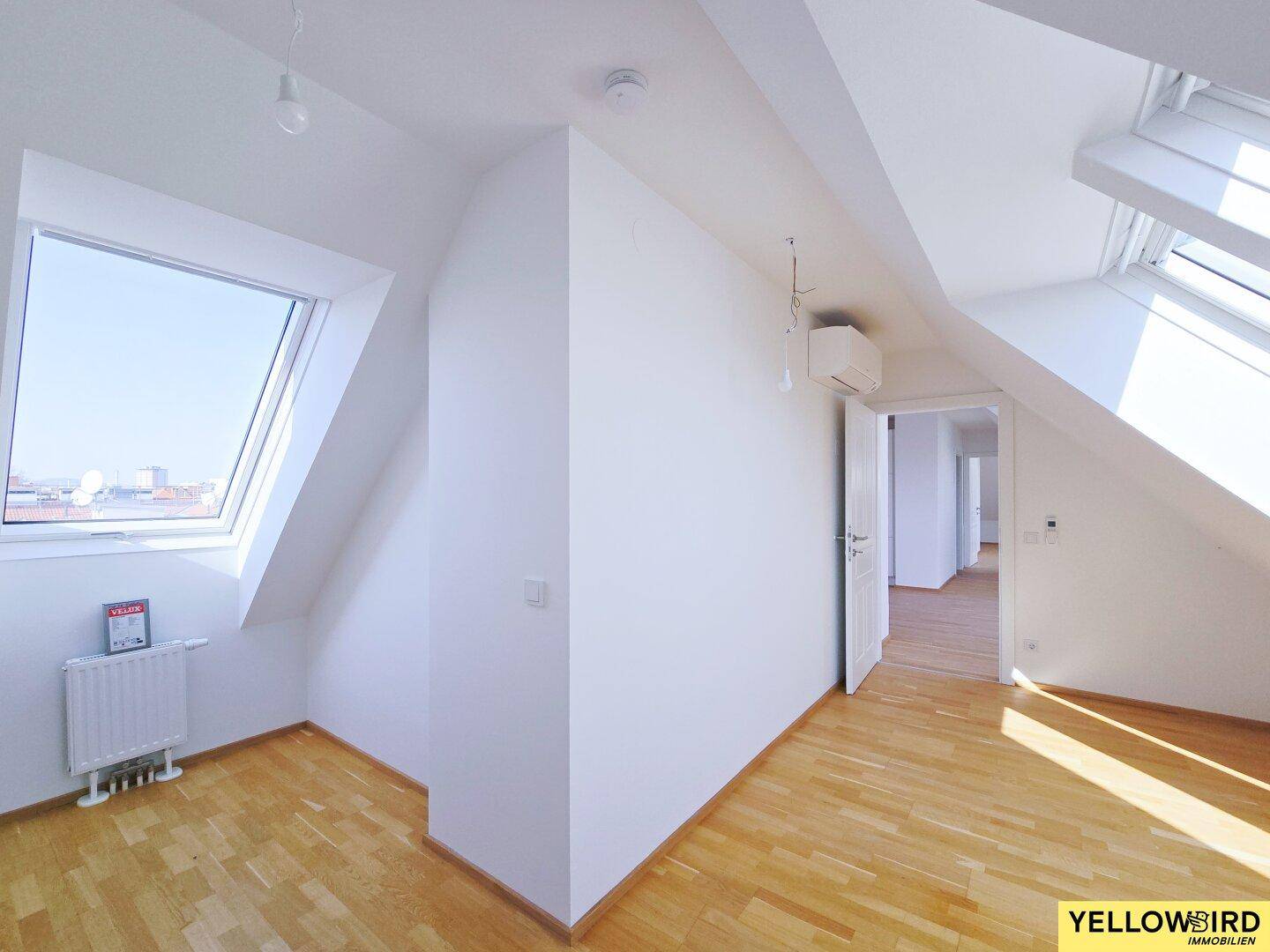 Heller Raum mit Holzboden, Dachfenster und Klimaanlage, mit Zugang zu weiteren Räumen.