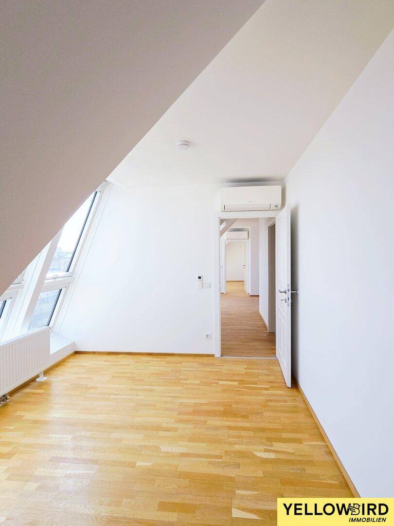 Heller Raum mit Holzboden, großer Fensterfront und Klimaanlage, mit Zugang zu einem weiteren Zimmer.