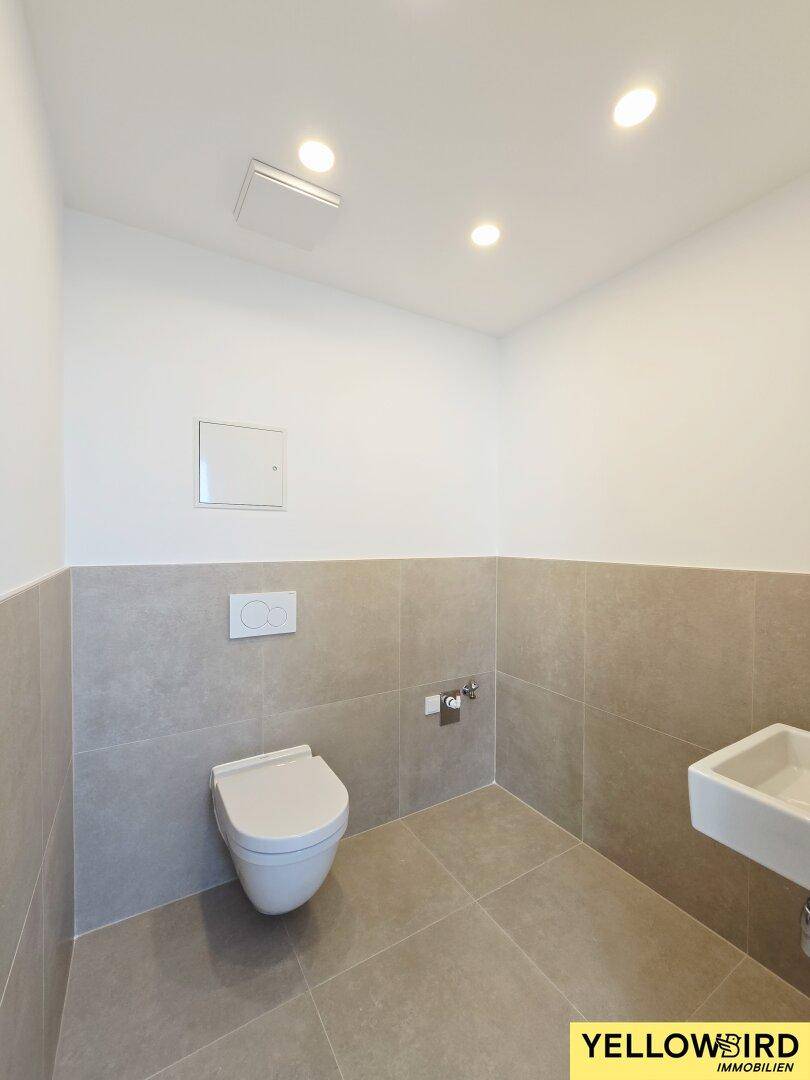 Separates WC mit modernen grauen Fliesen, wandhängender Toilette und kleinem Waschbecken.