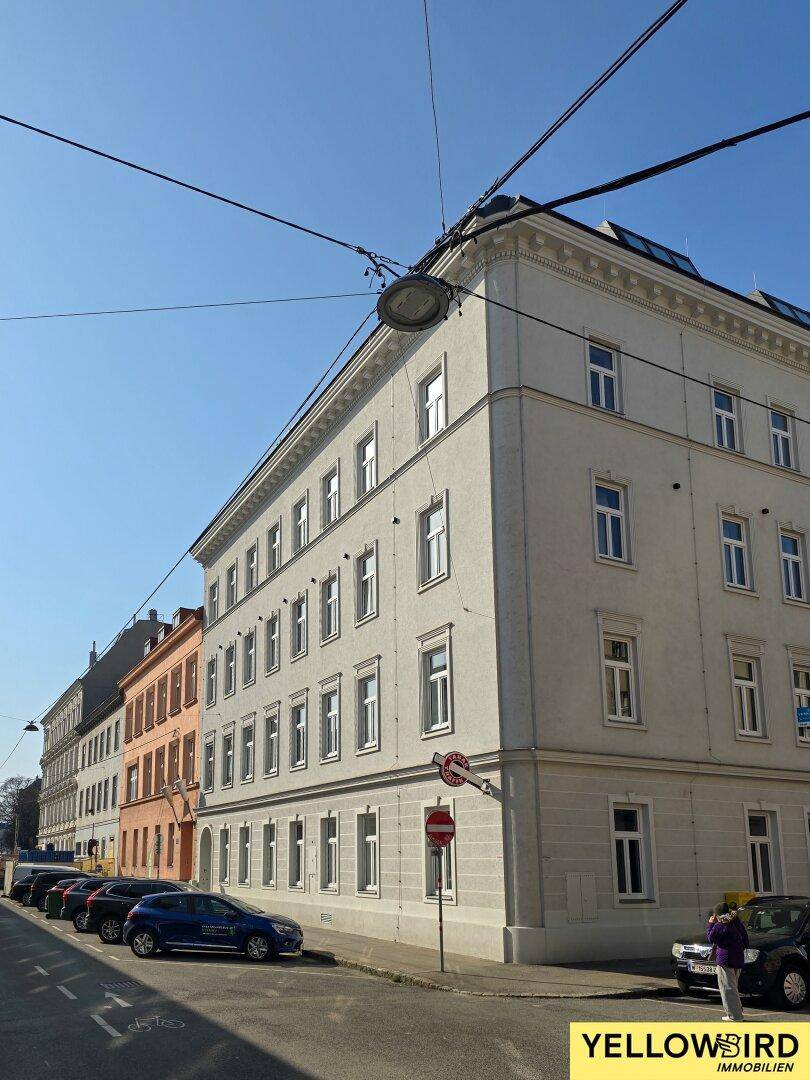 Historisches Eckgebäude an einer belebten Straße mit parkenden Autos und klarem Himmel.