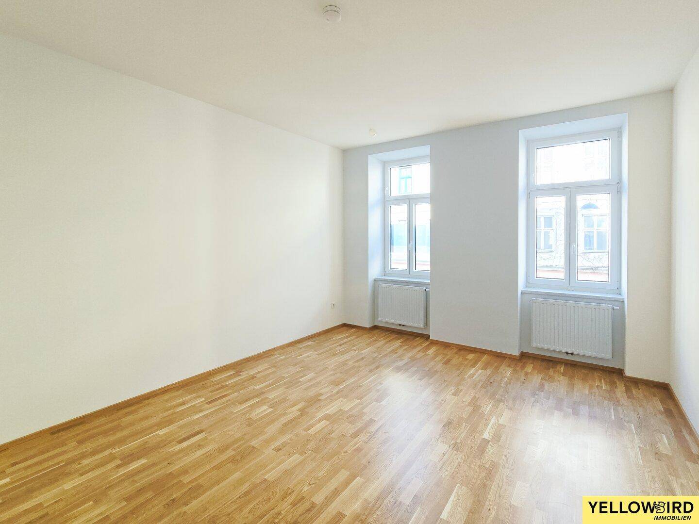 Geräumiges, leeres Zimmer mit hellem Holzboden und zwei großen Fenstern, bereit zur individuellen Gestaltung.