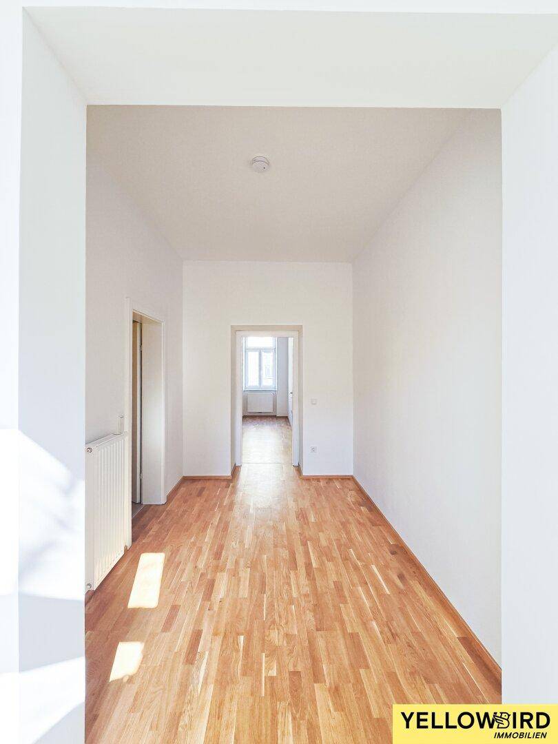 Langer, heller Flur mit Holzboden, der zu einem Zimmer mit Fenster am Ende führt.