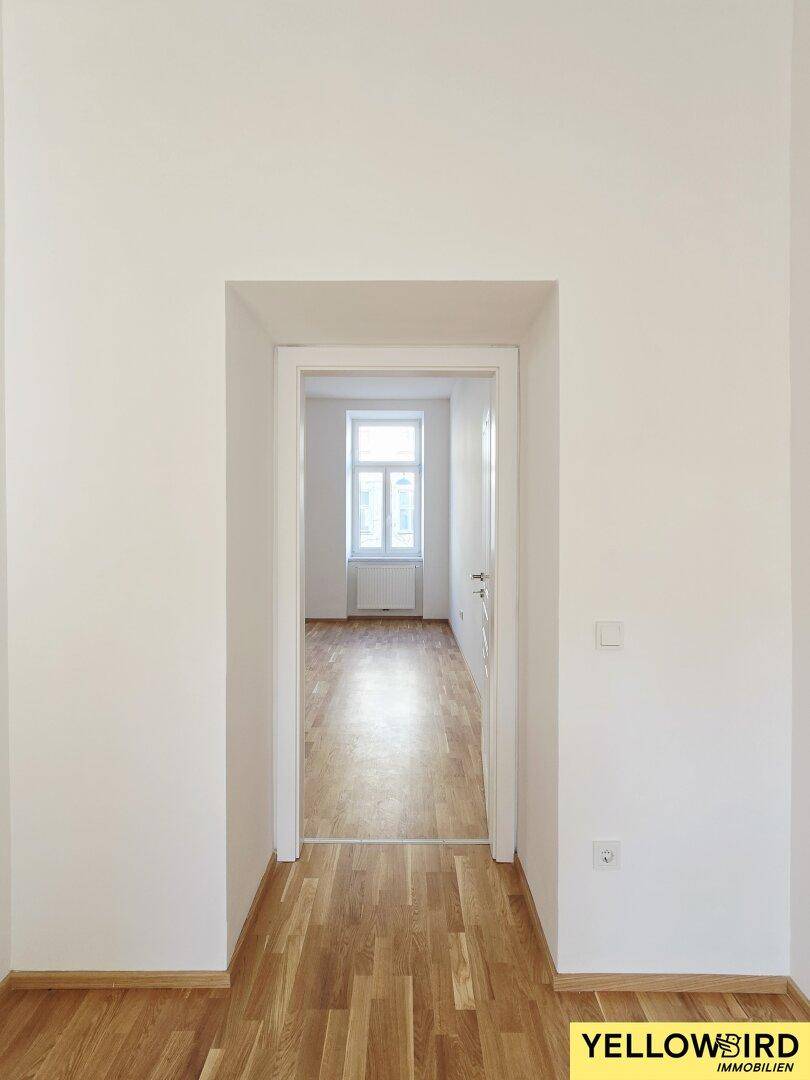 Heller Türrahmen öffnet sich zu einem leeren Zimmer mit Holzboden und einem Fenster.