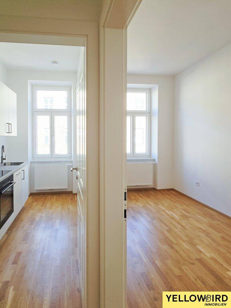 Blick von einem leeren Raum in eine moderne, weiße Küche mit Fenster.