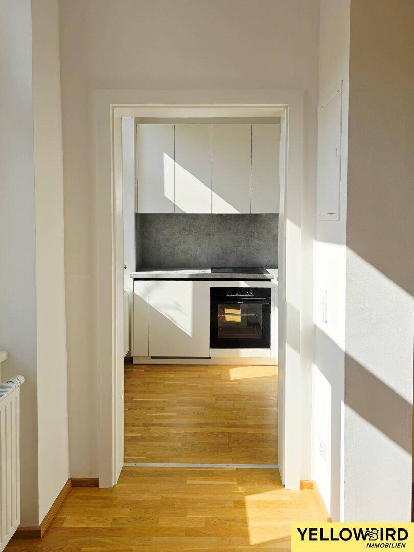 Blick durch einen Türrahmen in eine moderne Küche mit weißen Schränken und dunkler Arbeitsplatte.
