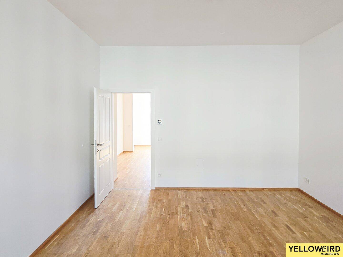 Leerer Raum mit Holzboden und weiß gestrichenen Wänden, mit offener Tür zu einem weiteren Zimmer.