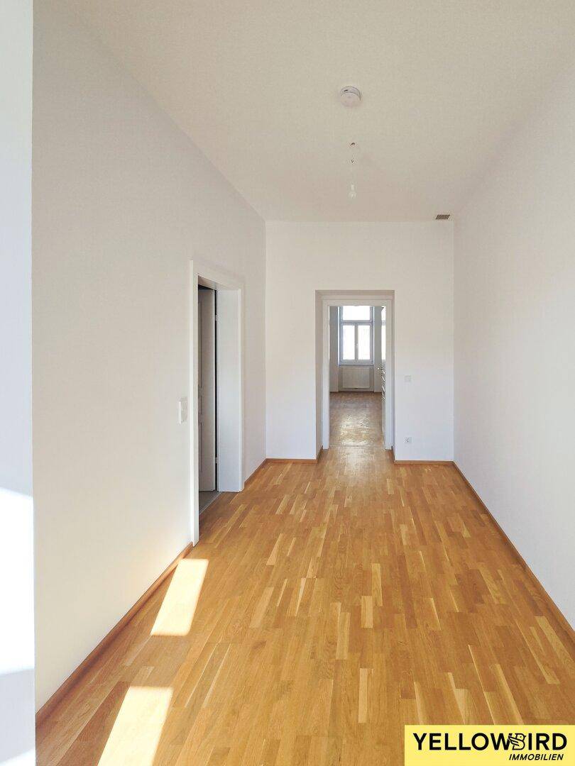 Leerer Flur mit hellem Holzboden und weißen Wänden, der zu einem weiteren Raum führt.