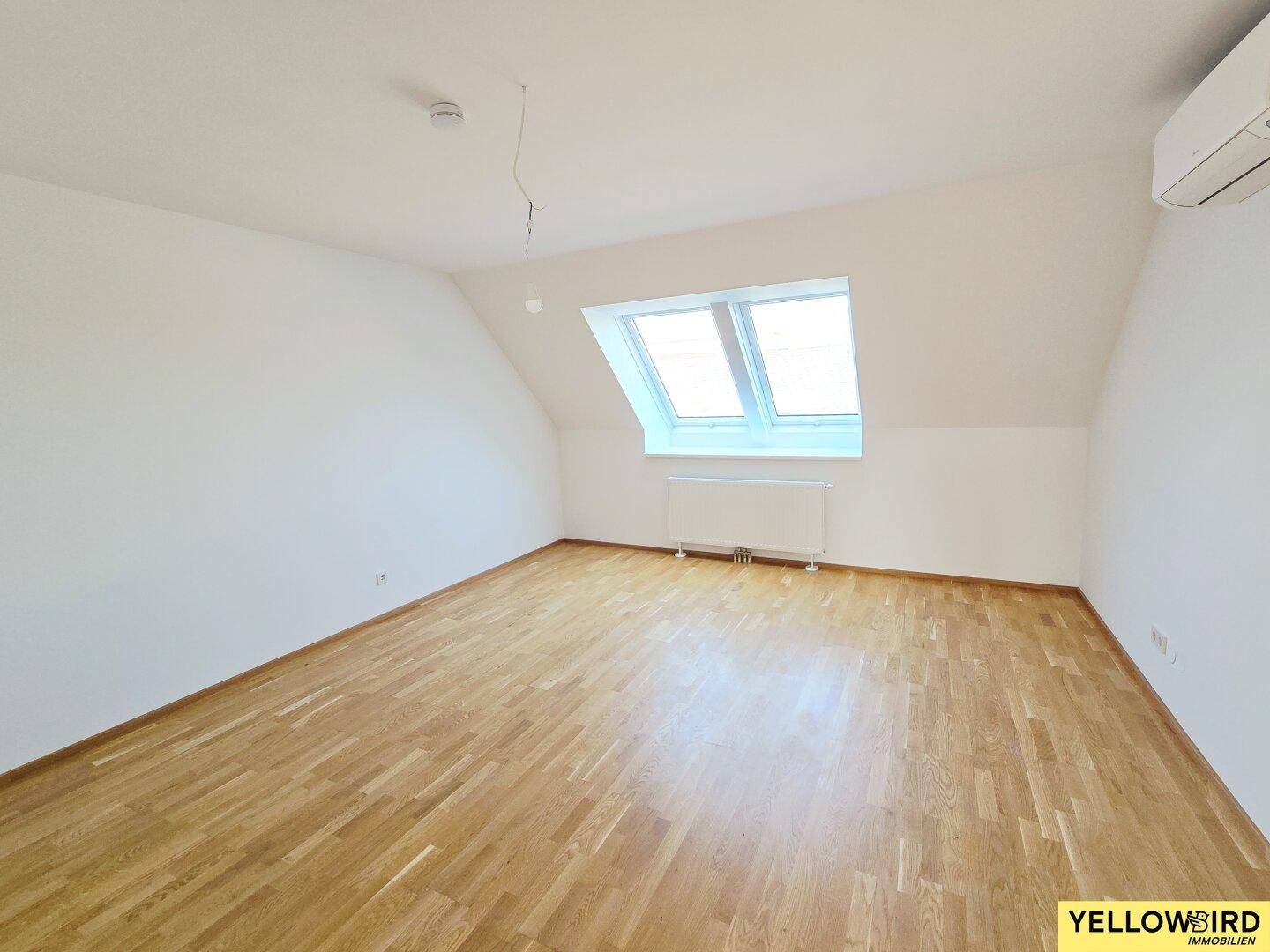 Helles Zimmer im Dachgeschoss mit Holzboden, Dachfenstern und Klimaanlage.