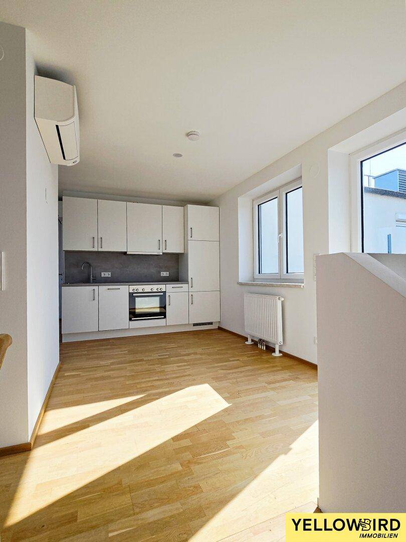 Helle, leere Küche mit weißen Schränken, Holzboden und Heizkörper unter dem Fenster.