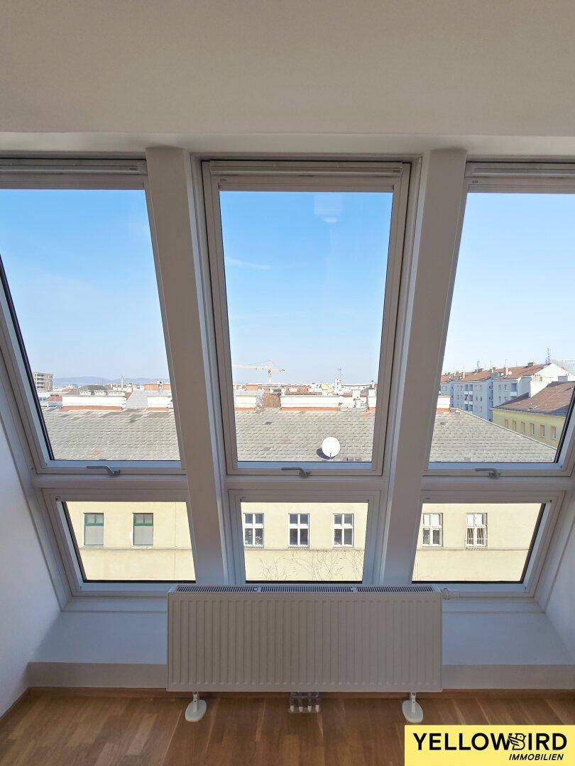 Heller Dachgeschossraum mit großen Fenstern, Holzboden und Blick auf die Stadt.
